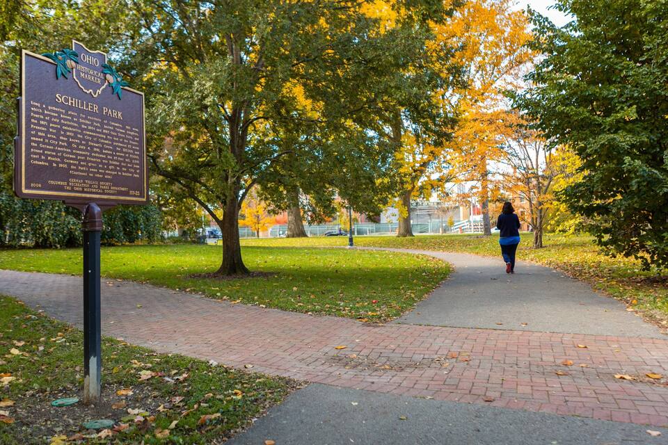 A park with a historical marker, trees with autumn leaves, and a paved path.