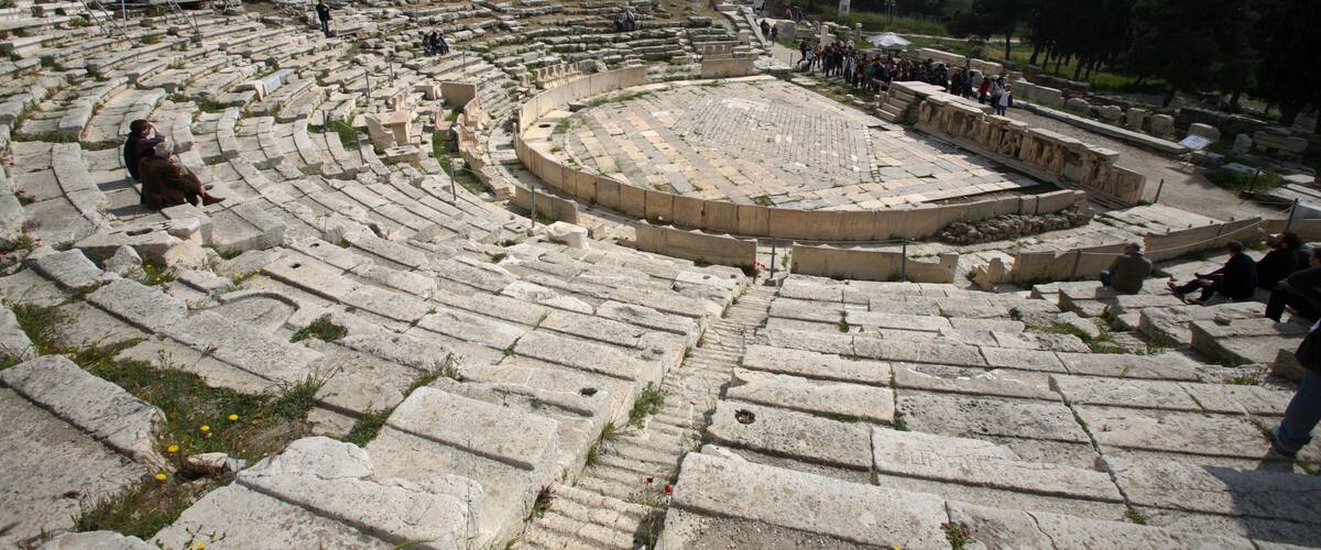 An ancient amphitheater with stone seating and a central stage, surrounded by trees and a cityscape in the background.