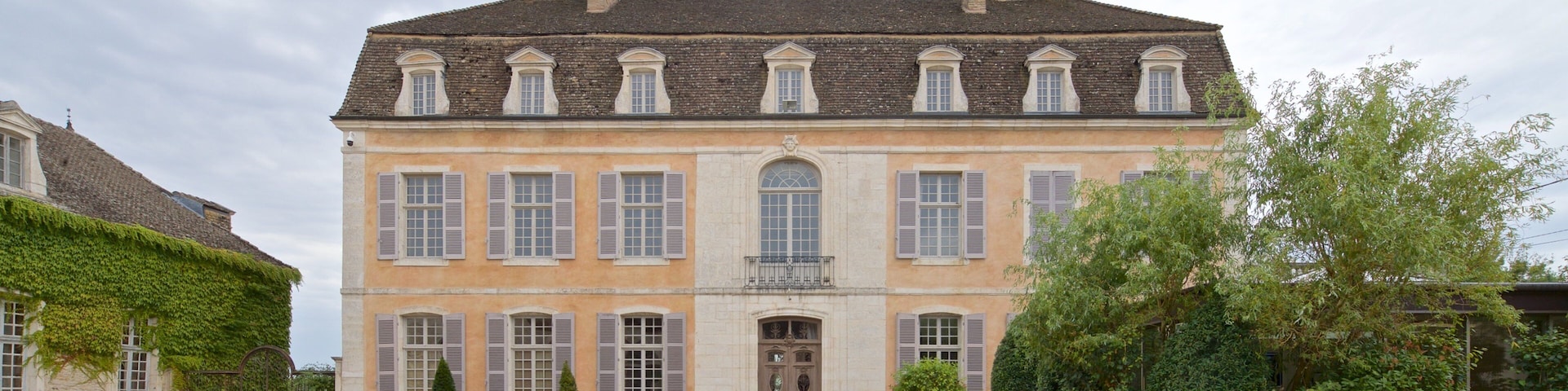 A large, historic building with a symmetrical facade, a central entrance, and a formal garden with geometrically shaped hedges.
