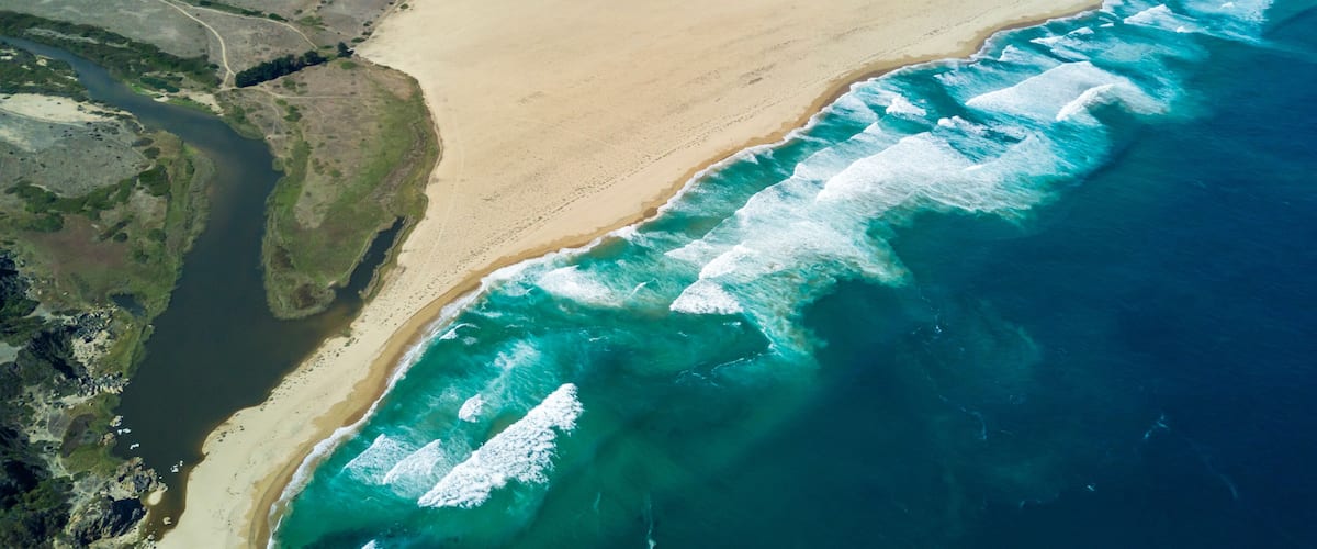A coastal landscape with a sandy beach, ocean waves, and a mountainous backdrop.