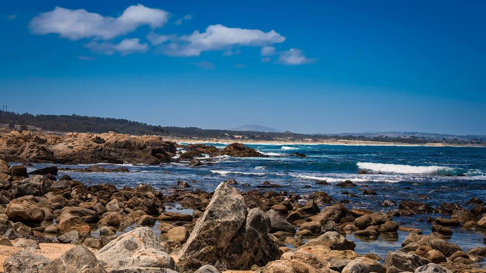 A rocky coastline with waves crashing against the shore under a clear blue sky.