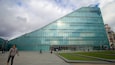 A modern glass building with a large glass facade, surrounded by a plaza with grass and a few people walking.