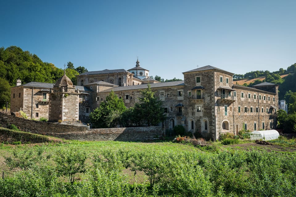 A historic stone building with a bell tower, surrounded by lush greenery and vineyards.