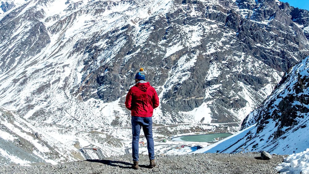 A person standing on a rocky mountain edge, overlooking a valley with snow-covered slopes and a lake.
