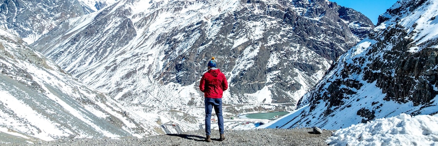 A person standing on a rocky mountain edge, overlooking a valley with snow-covered slopes and a lake.