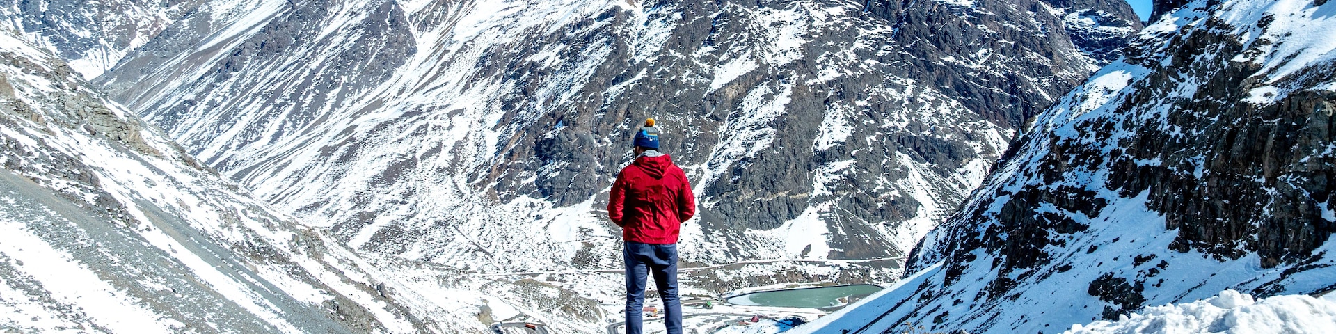 Una persona parada al borde de una montaña rocosa, contemplando un valle con laderas nevadas y un lago.