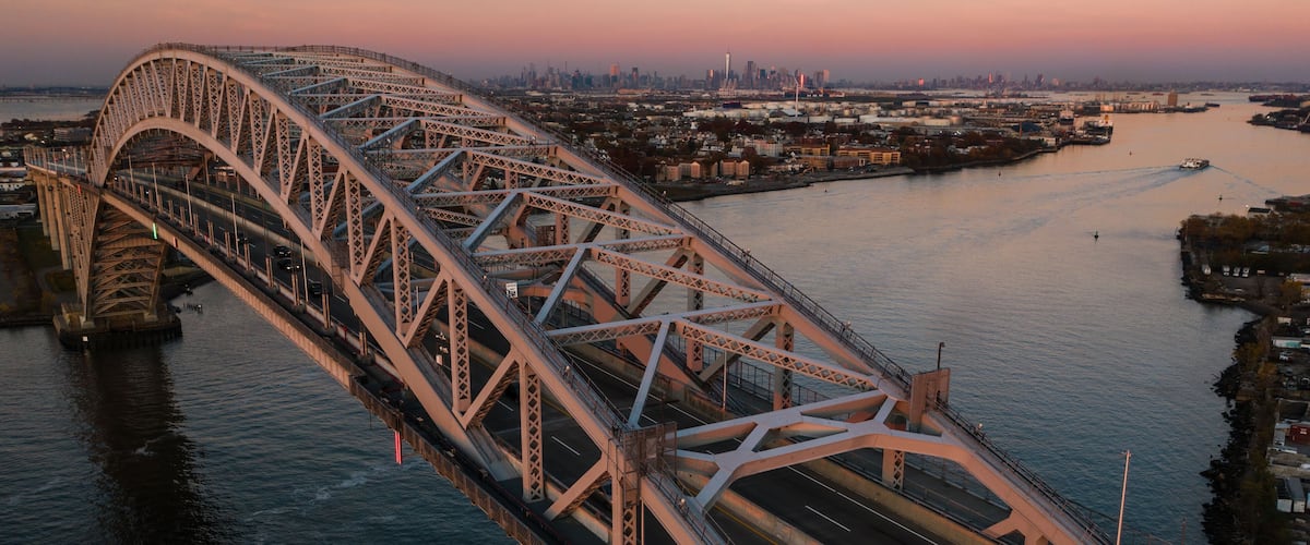 A large steel bridge spanning a wide river, with a city skyline in the background.