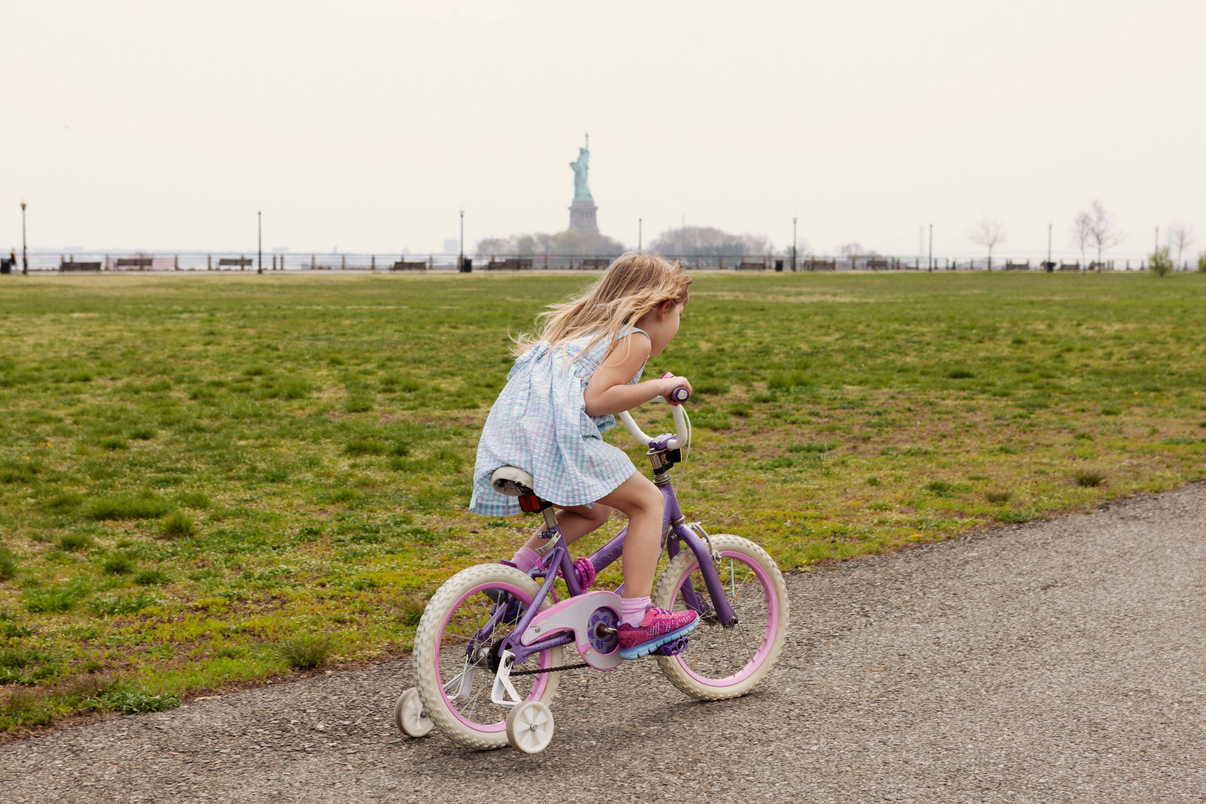 Side view of girl riding bicycle on road by field with Statue of Liberty in background