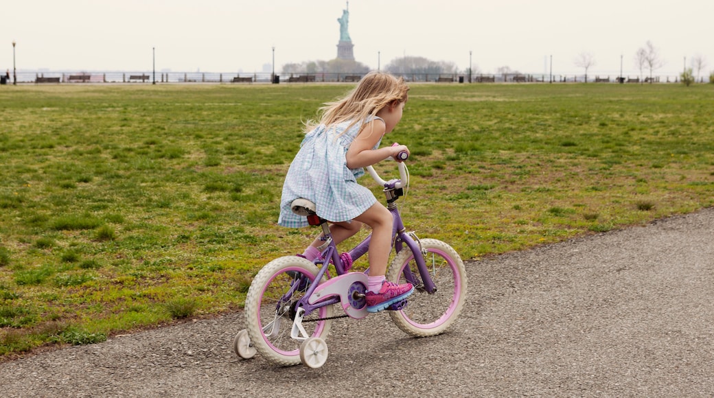 Side view of girl riding bicycle on road by field with Statue of Liberty in background