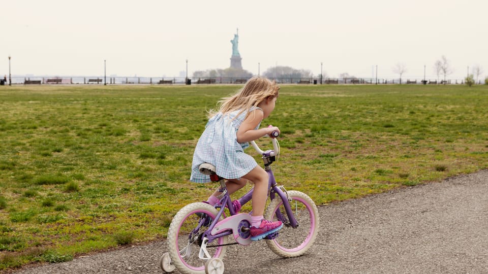 A child riding a purple bicycle on a path with the Statue of Liberty in the background.