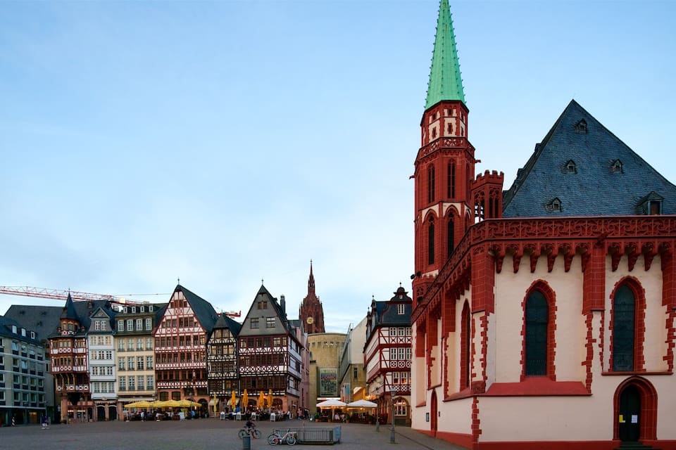 A historic European town square with traditional half-timbered buildings, a church with a tall spire, and a modern building with a crane.