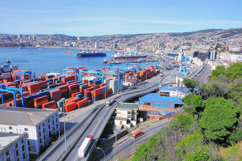 Un puerto con contenedores de carga, barcos y un paisaje urbano al fondo.