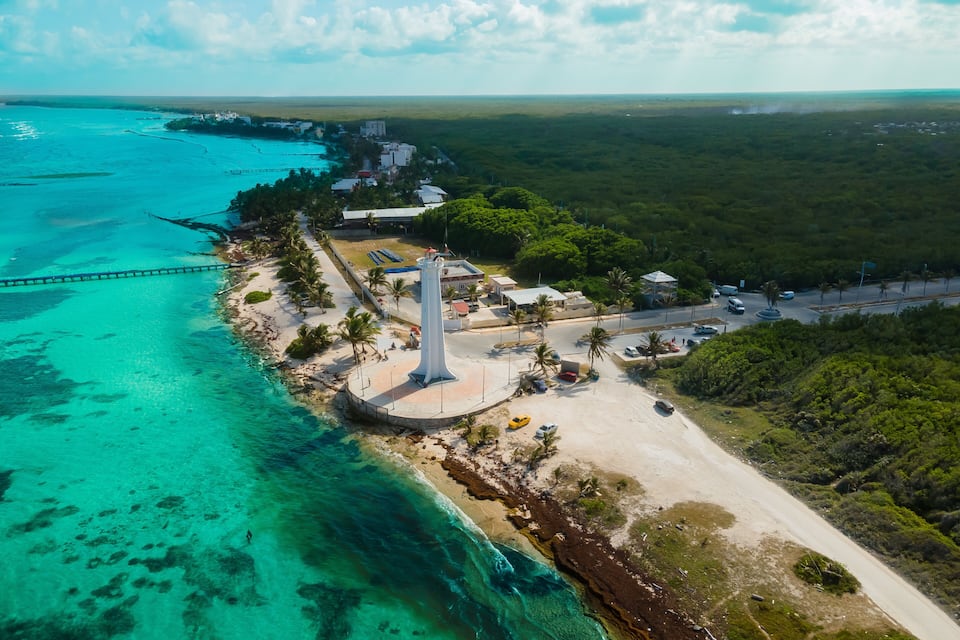 A coastal area with a lighthouse, beach, clear turquoise waters, and a bridge.