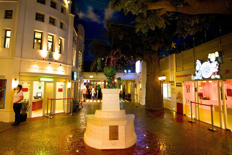 Ein beleuchteter Stadtplatz bei Nacht mit einem zentralen Brunnen, umgebenden Gebäuden und einem Baum.