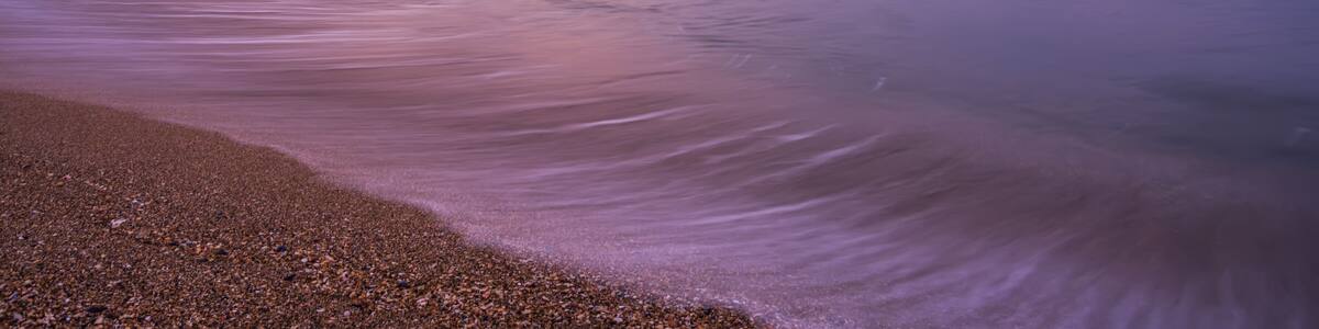 A pier extending into calm waters with a pebbly beach in the foreground and a purple sky.