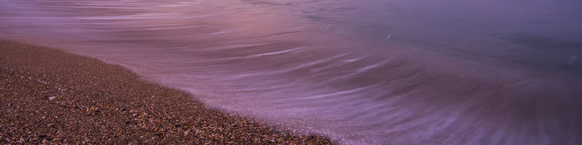 A pier extending into calm waters with a pebbly beach in the foreground and a purple sky.