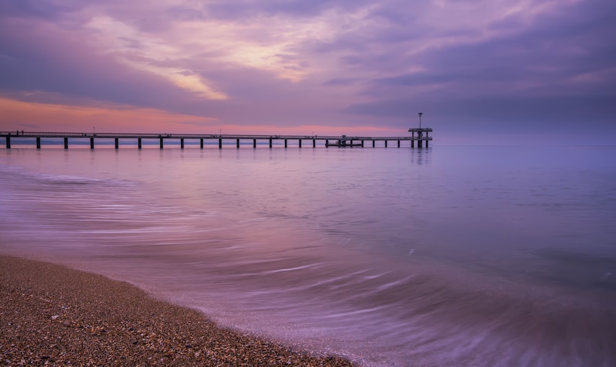A pier extending into calm waters with a pebbly beach in the foreground and a purple sky.