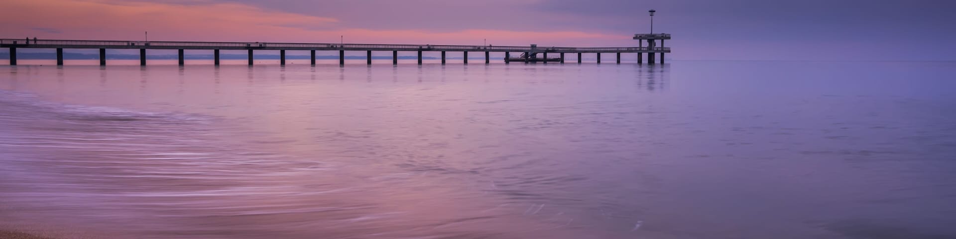 Un muelle se adentra en aguas tranquilas, con una playa de guijarros en primer plano y un cielo morado.