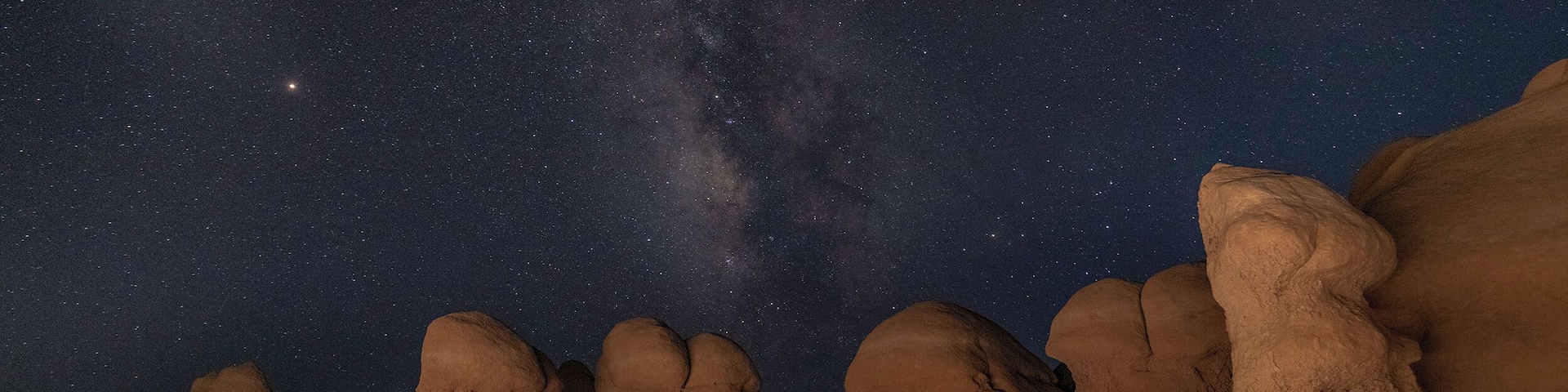 Céu noturno repleto de estrelas e da Via Láctea sobre uma paisagem rochosa com formações singulares.