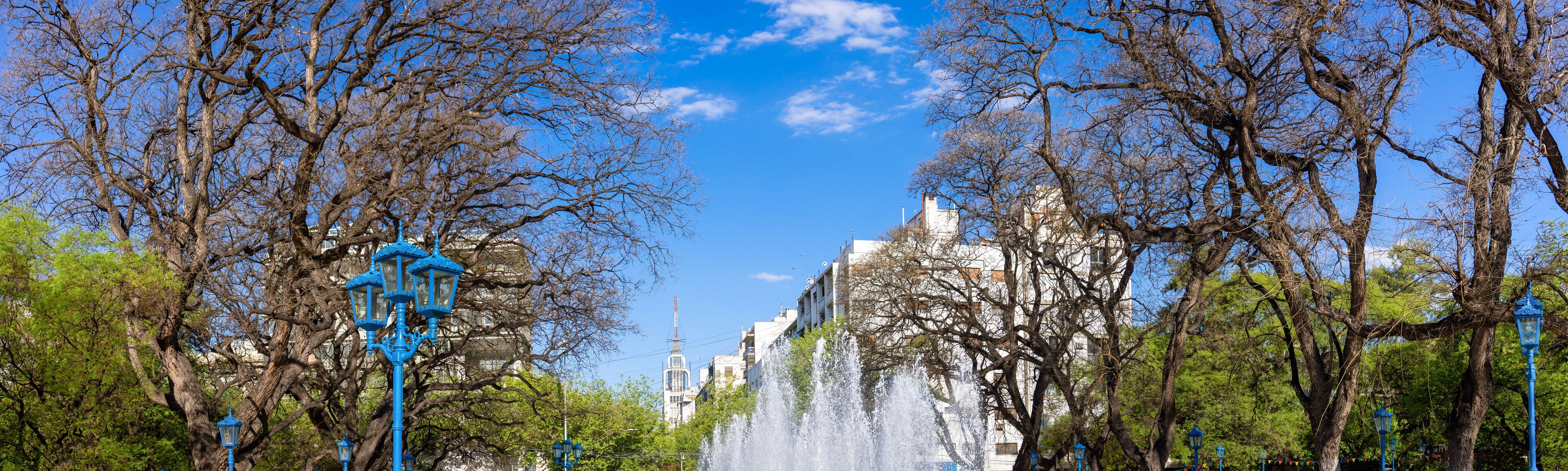 Um parque com uma fonte, árvores e um edifício ao fundo.