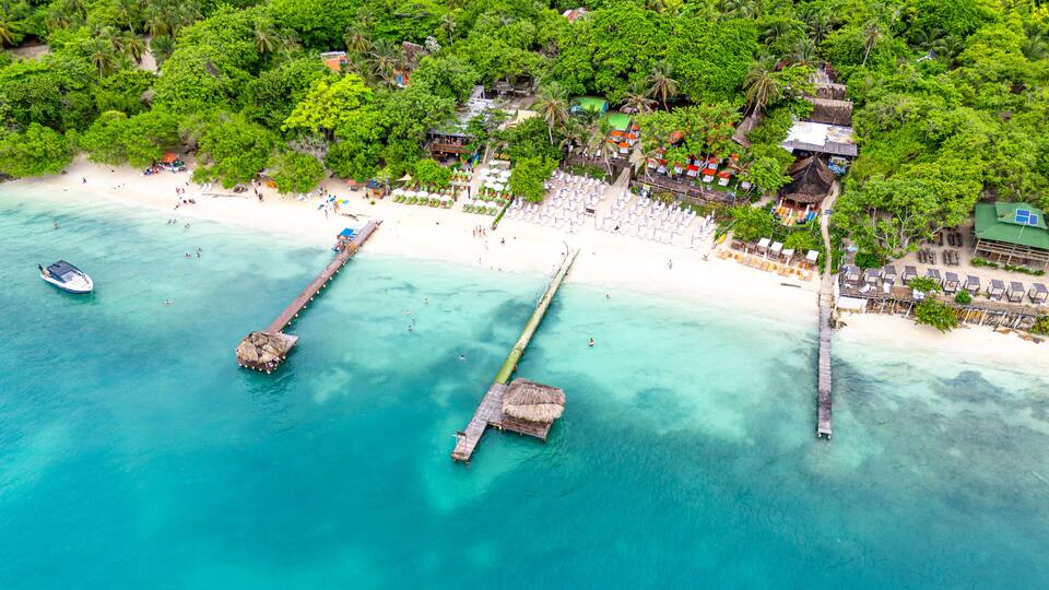 A tropical beach with clear turquoise waters, a pier, and a resort area with umbrellas and beach chairs.