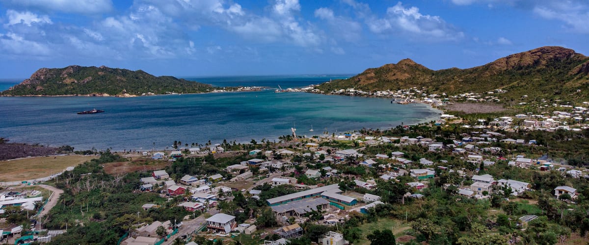 A coastal town with a clear blue sea, a lighthouse, and mountains in the background.