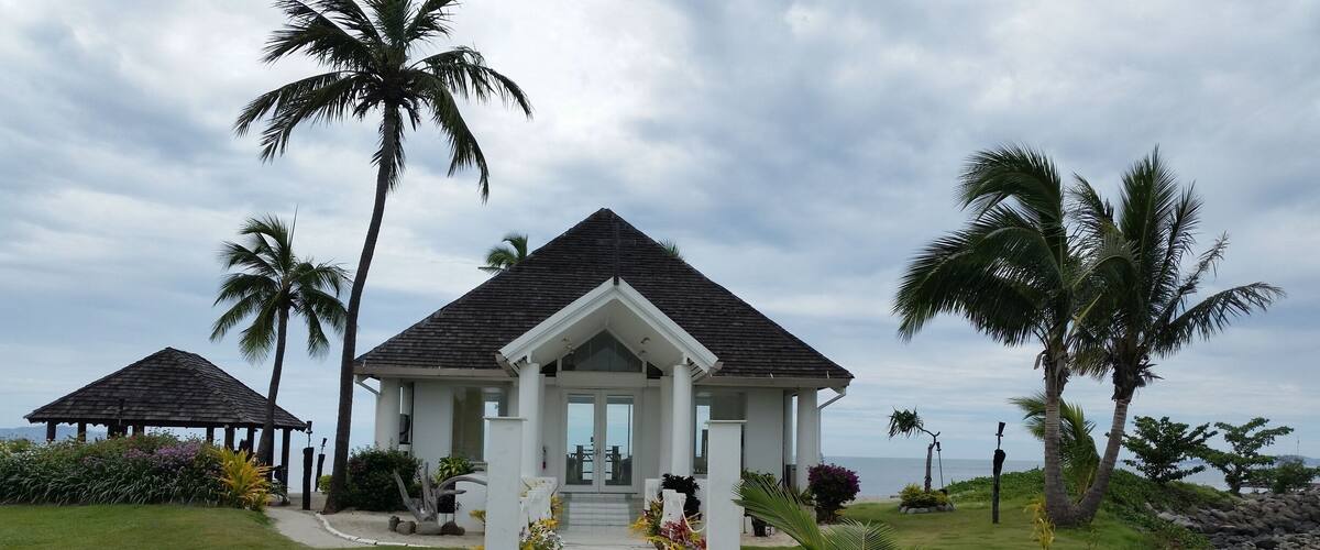 A white building with a triangular roof, surrounded by palm trees and a garden.