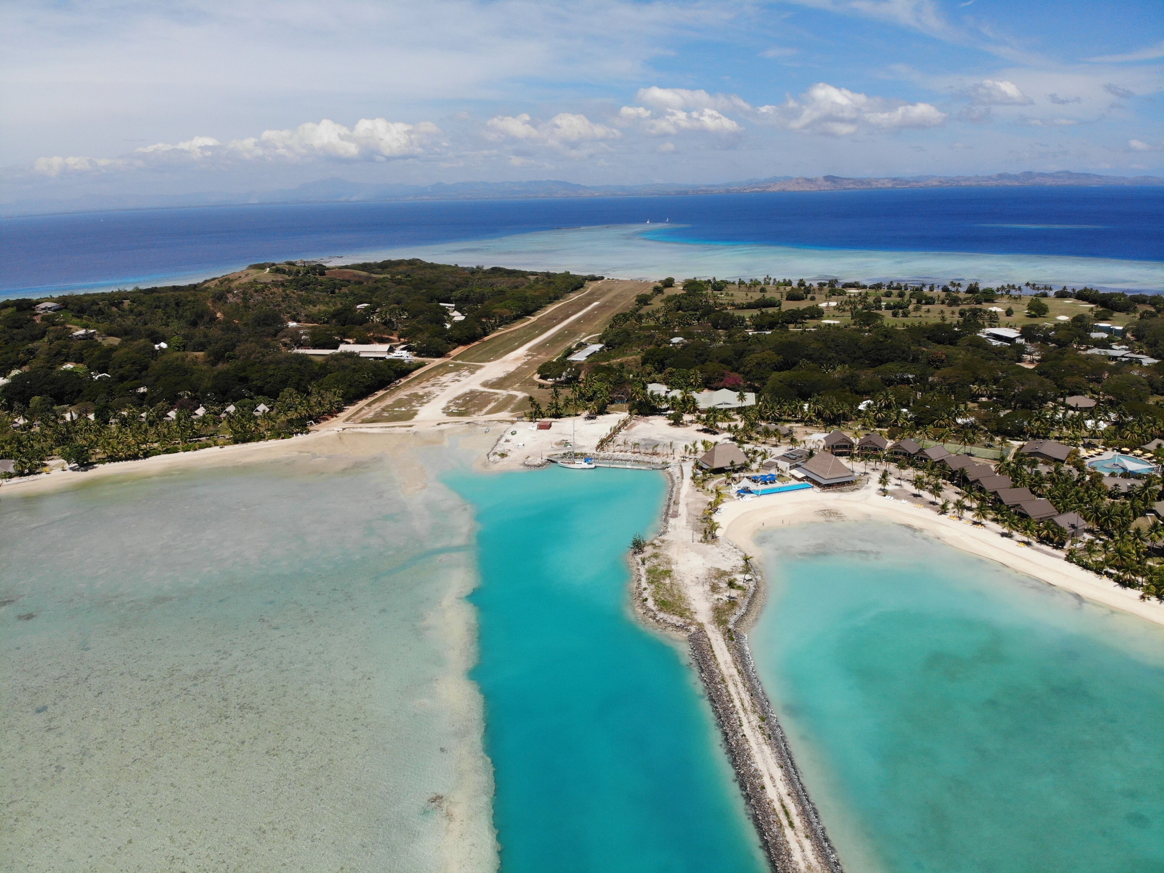 Vista aérea de una zona costera con aguas turquesas cristalinas, una playa de arena y una combinación de edificios residenciales y vegetación.