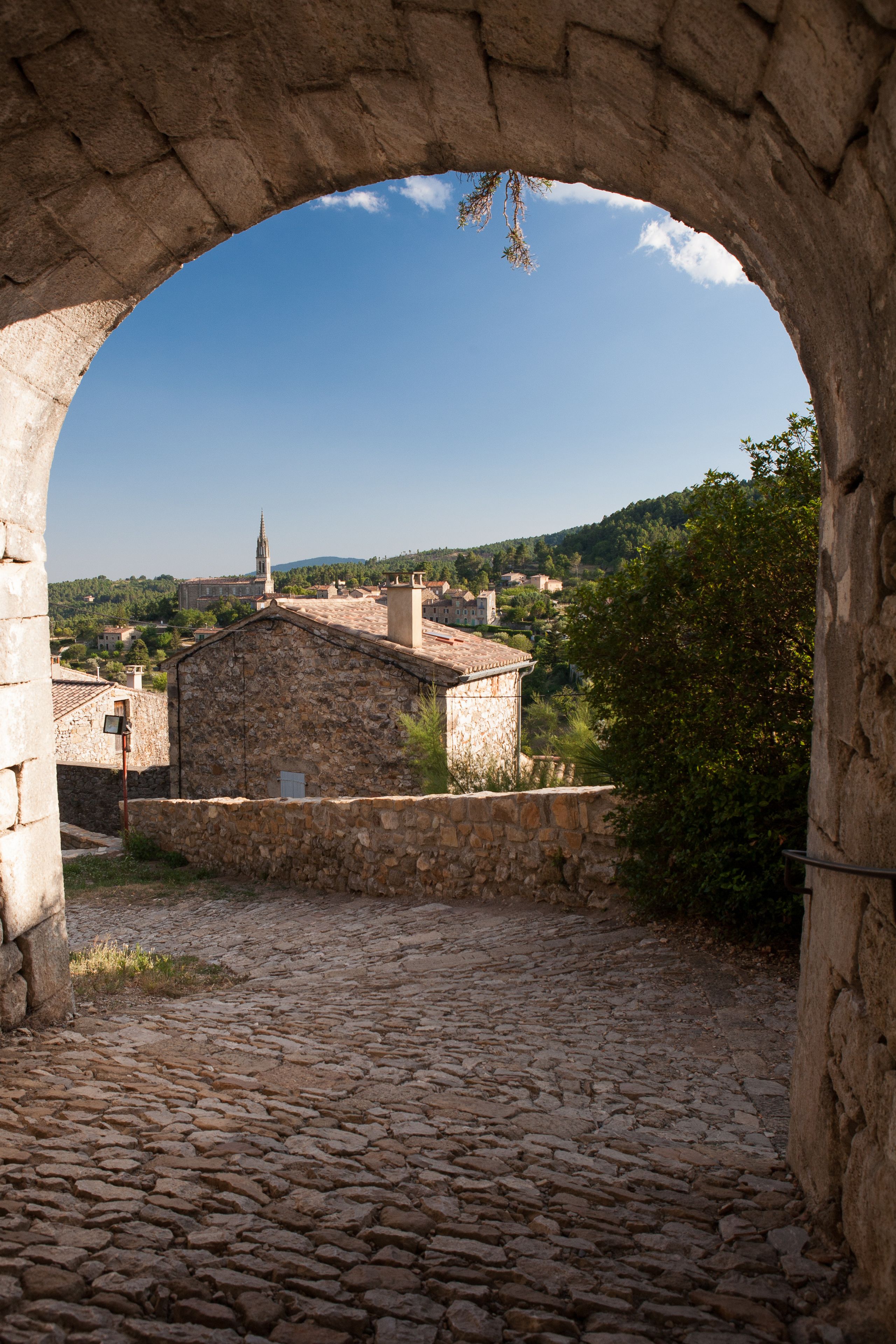 An arched stone structure with a cobblestone courtyard, a church with a bell tower, and a hilly landscape in the background.