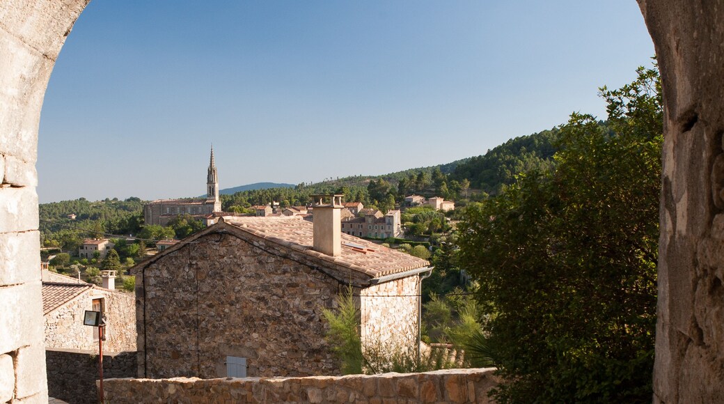 An arched stone structure with a cobblestone courtyard, a church with a bell tower, and a hilly landscape in the background.
