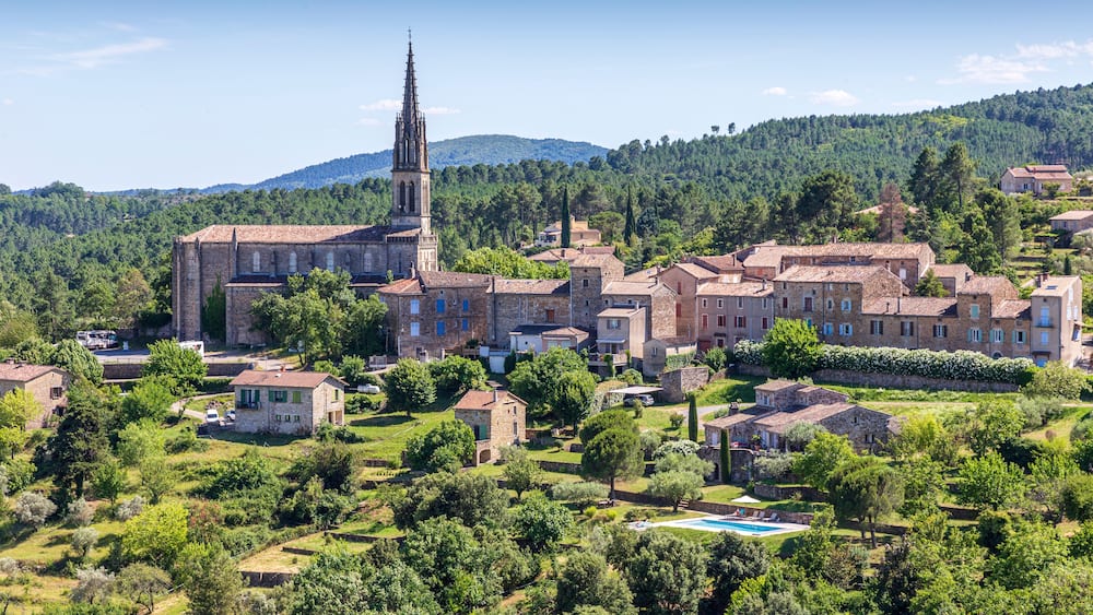 Une ville historique abritant une église remarquable, reconnaissable à son clocher imposant, et entourée d’une végétation luxuriante, avec en toile de fond un paysage montagneux.