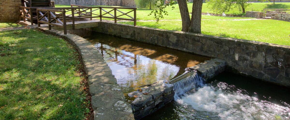 A water mill with a wooden bridge over a stream, surrounded by trees and grassy areas.