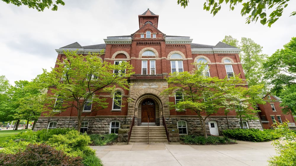 A large red brick building with a central arched entrance, flanked by stone columns, and surrounded by lush greenery.