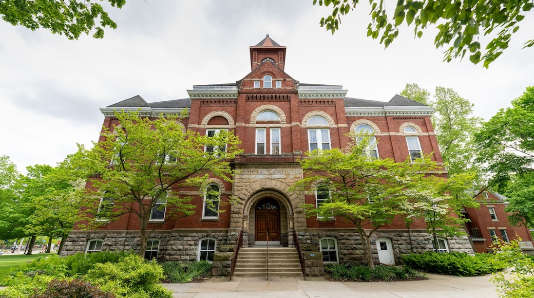 A large red brick building with a central arched entrance, flanked by stone columns, and surrounded by lush greenery.