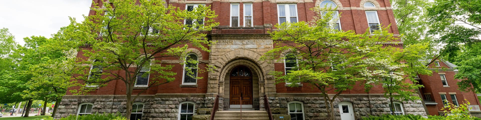 A large red brick building with a central arched entrance, flanked by stone columns, and surrounded by lush greenery.