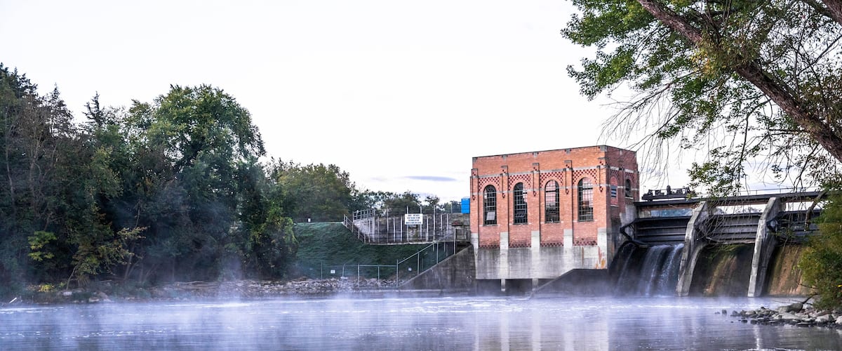 A river with a waterfall, a brick building, and surrounding greenery.
