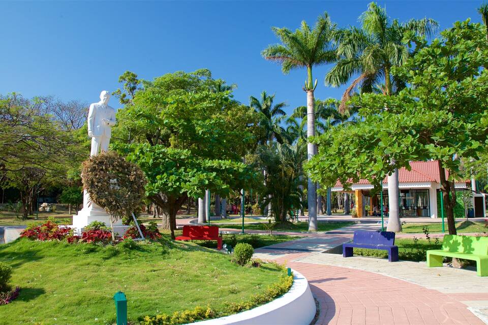 A statue in a park with palm trees, benches, and a building with a red roof.