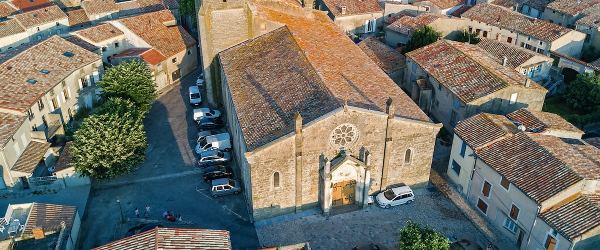 An aerial view of a historic town with tightly packed buildings, a prominent church with a rose window, and a narrow street with parked cars.