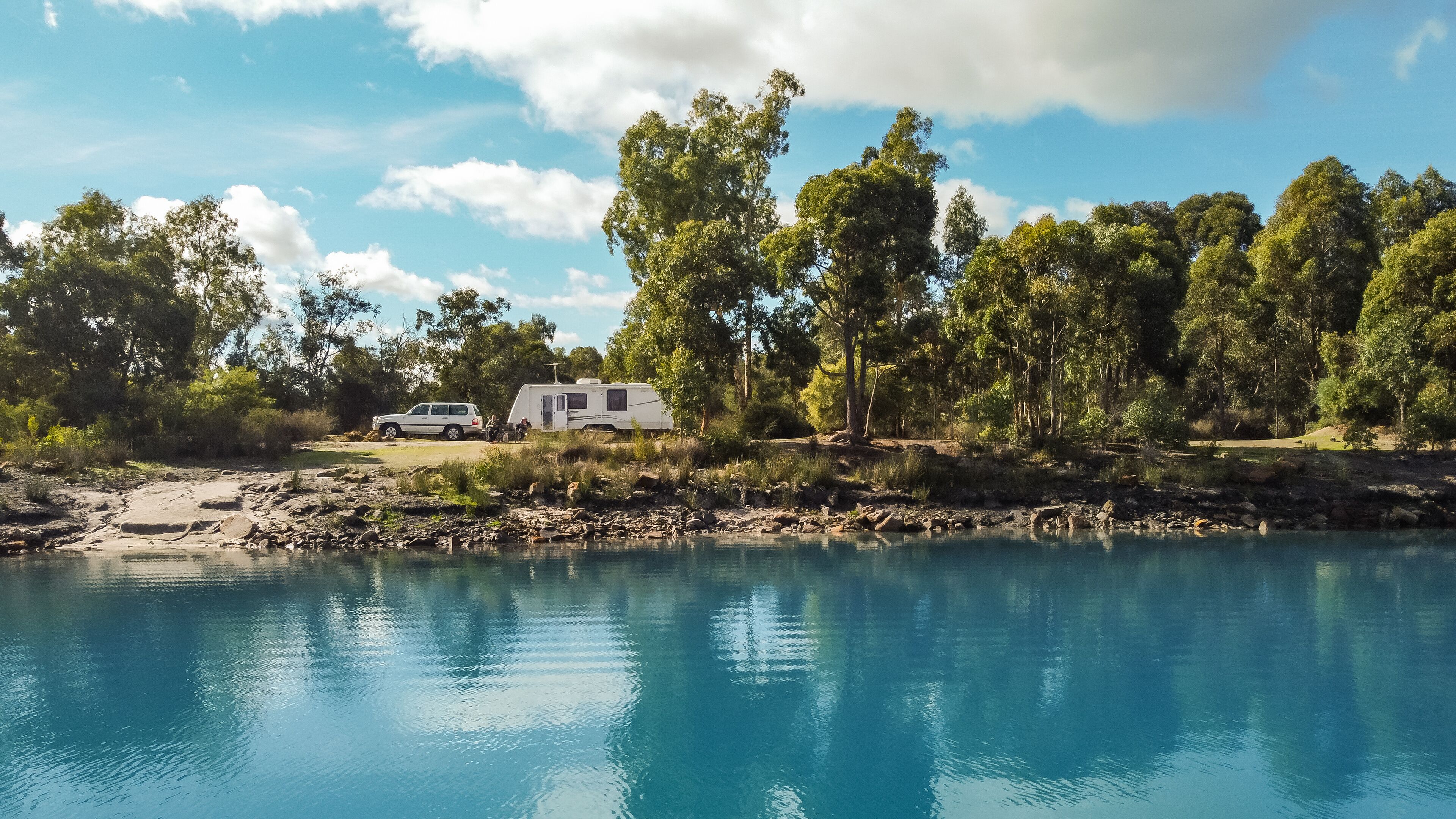 Landsscape view of a large modern caravan and four wheel drive vehicle free camping alongside a beautiful blue lake fringed with gumtrees.