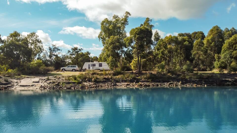 A serene lakeside campsite with a white RV and a trailer, surrounded by lush green trees and a clear blue sky.