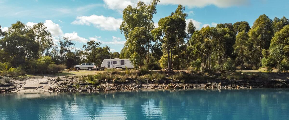 A serene lakeside campsite with a white RV and a trailer, surrounded by lush green trees and a clear blue sky.