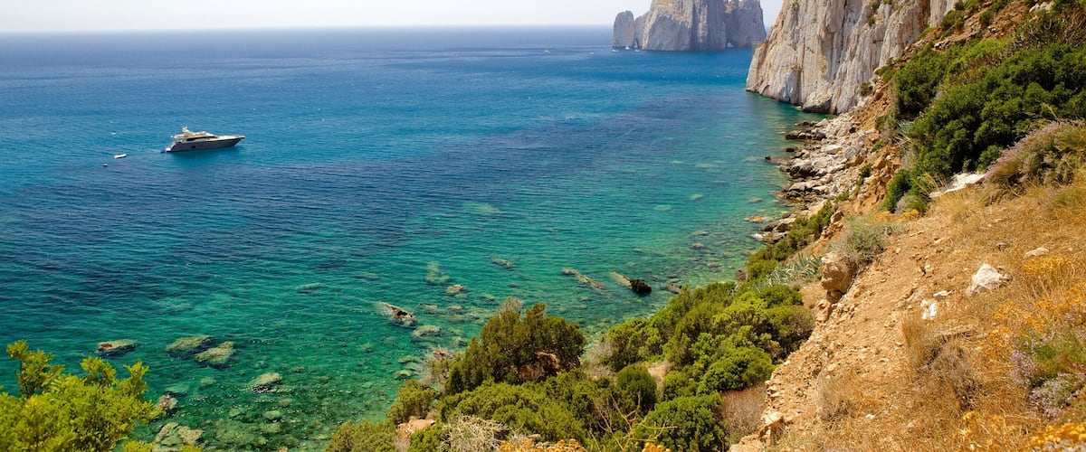 A coastal landscape with a yacht on clear turquoise waters, rocky cliffs, and lush greenery.