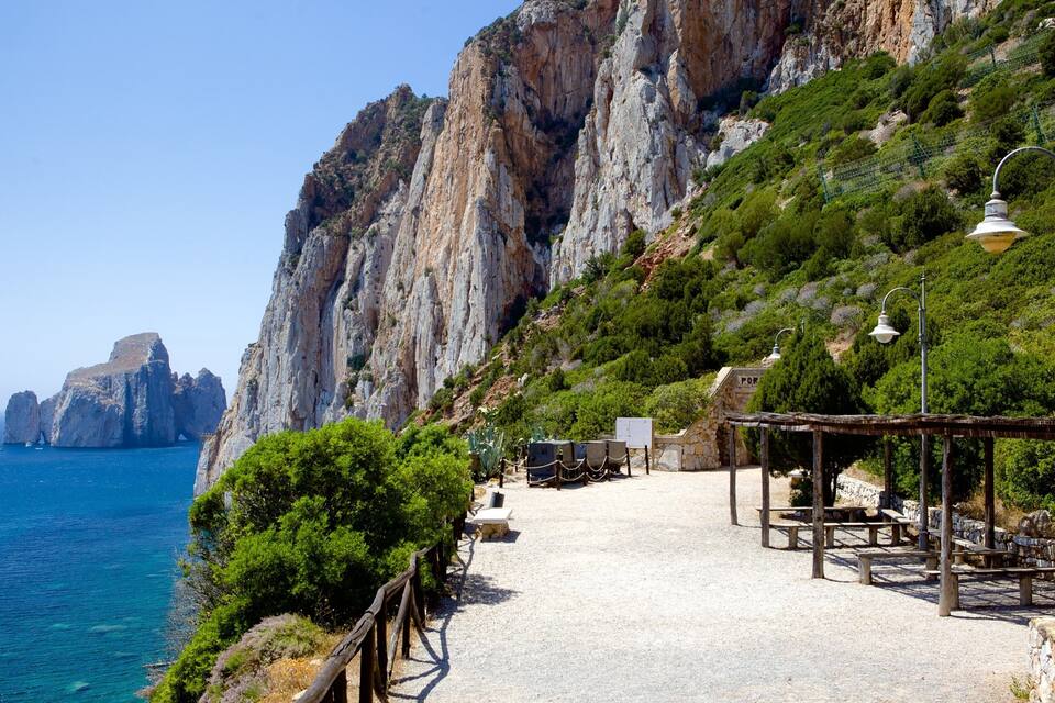Un sentier côtier bordé de falaises rocheuses, une mer d’un bleu limpide et une petite construction dotée de bancs.