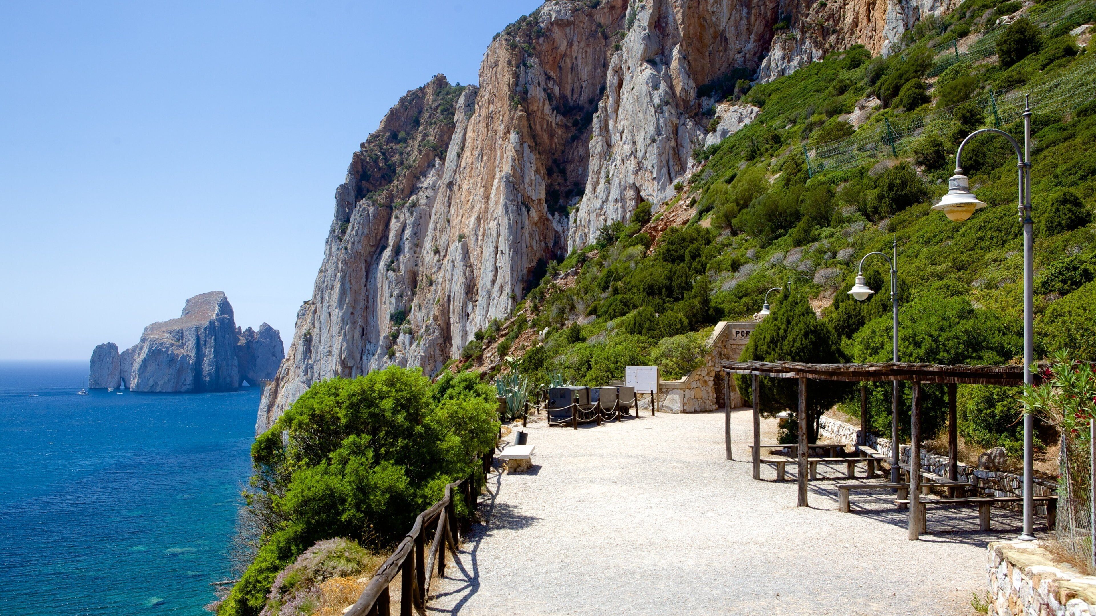 A coastal path with rocky cliffs, a clear blue sea, and a small structure with benches.