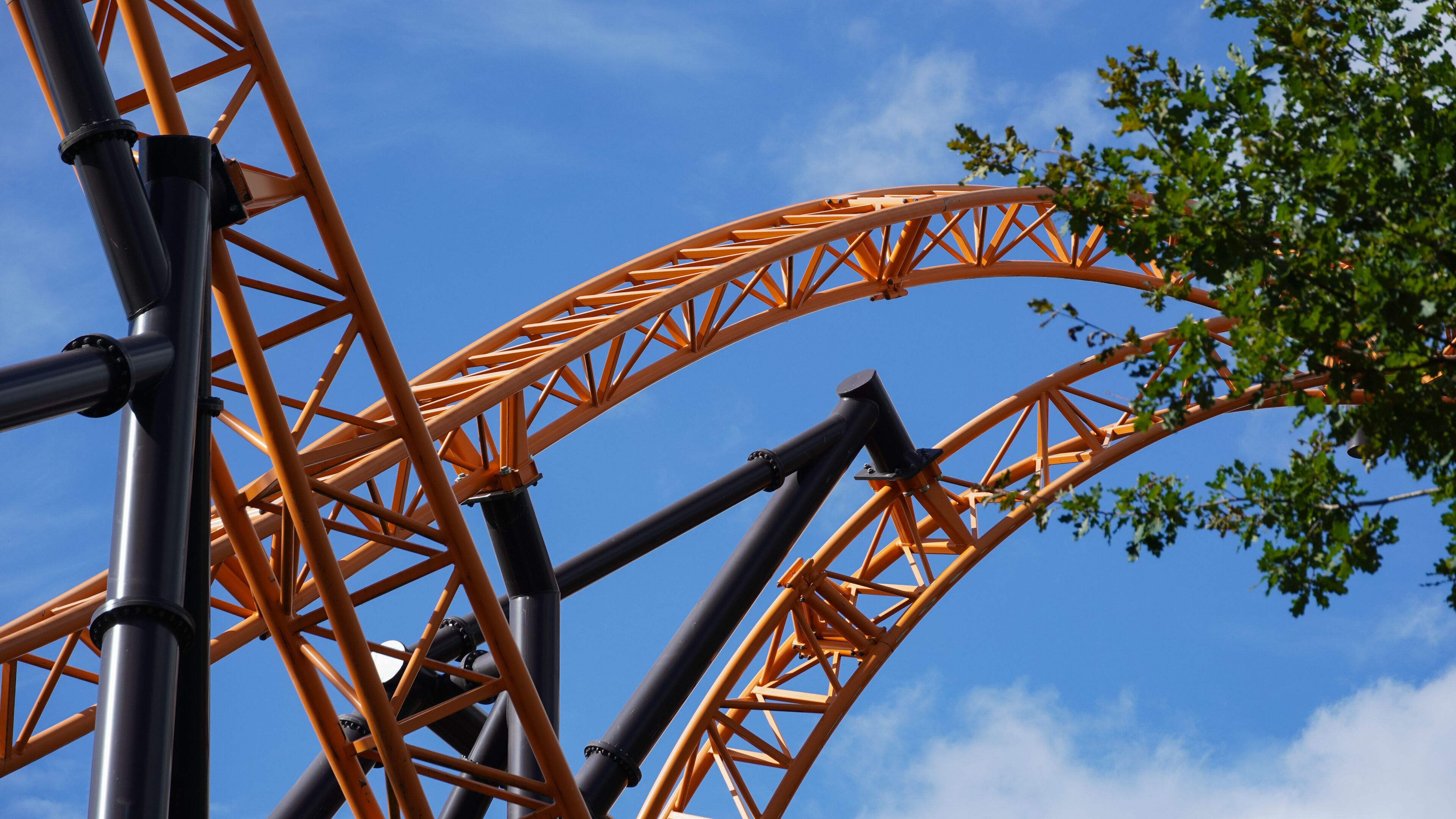 Orange-coloured roller coaster tracks of an extreme roller coaster, crossing under a blue sky and a tree in the foreground