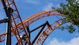 Orange-coloured roller coaster tracks of an extreme roller coaster, crossing under a blue sky and a tree in the foreground