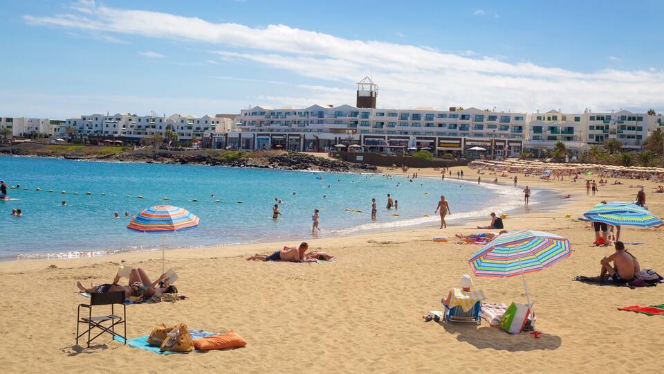 A beach with people swimming and sunbathing, umbrellas, and buildings in the background.