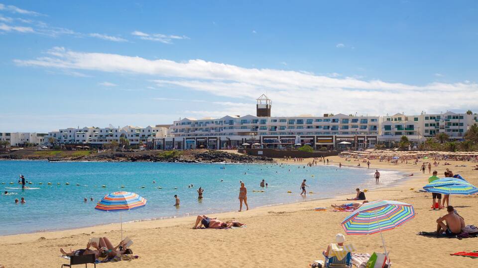 A beach with people swimming and sunbathing, umbrellas, and buildings in the background.