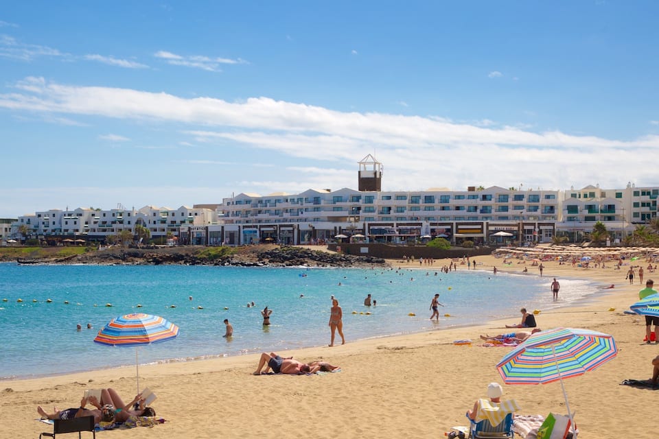 A beach with people swimming and sunbathing, umbrellas, and buildings in the background.