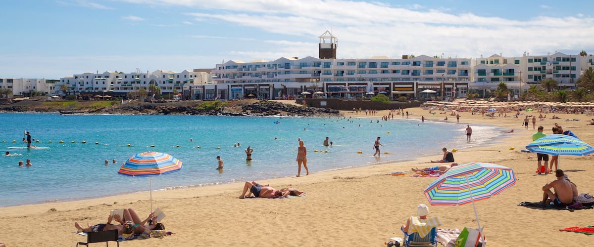 A beach with people swimming and sunbathing, umbrellas, and buildings in the background.